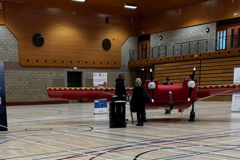 A small red plane on display in a large gymnasium hall with two people standing next to it